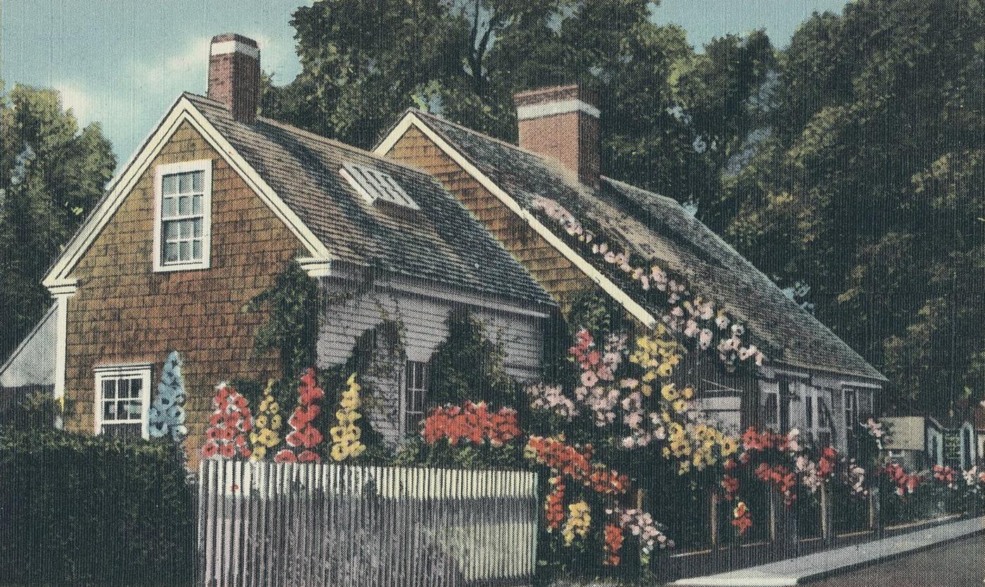 Postcard: a weathered shingle cape house with two chimneys, hollyhocks and roses along a white picket fence
