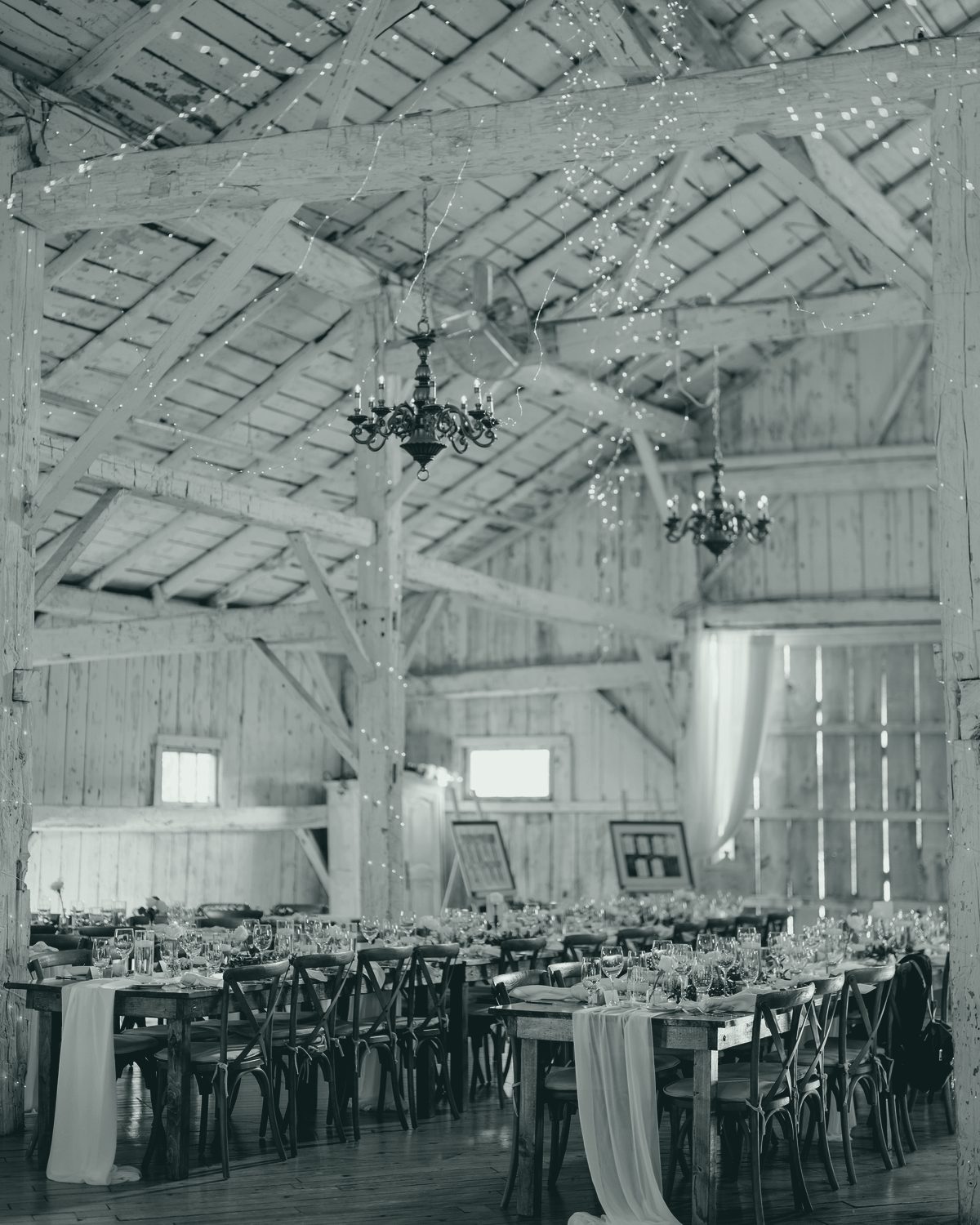 Inside the hall: whitewashed timber trusses and plank walls, two brass chandeliers, long wooden tables set for dinner with cross-back chairs