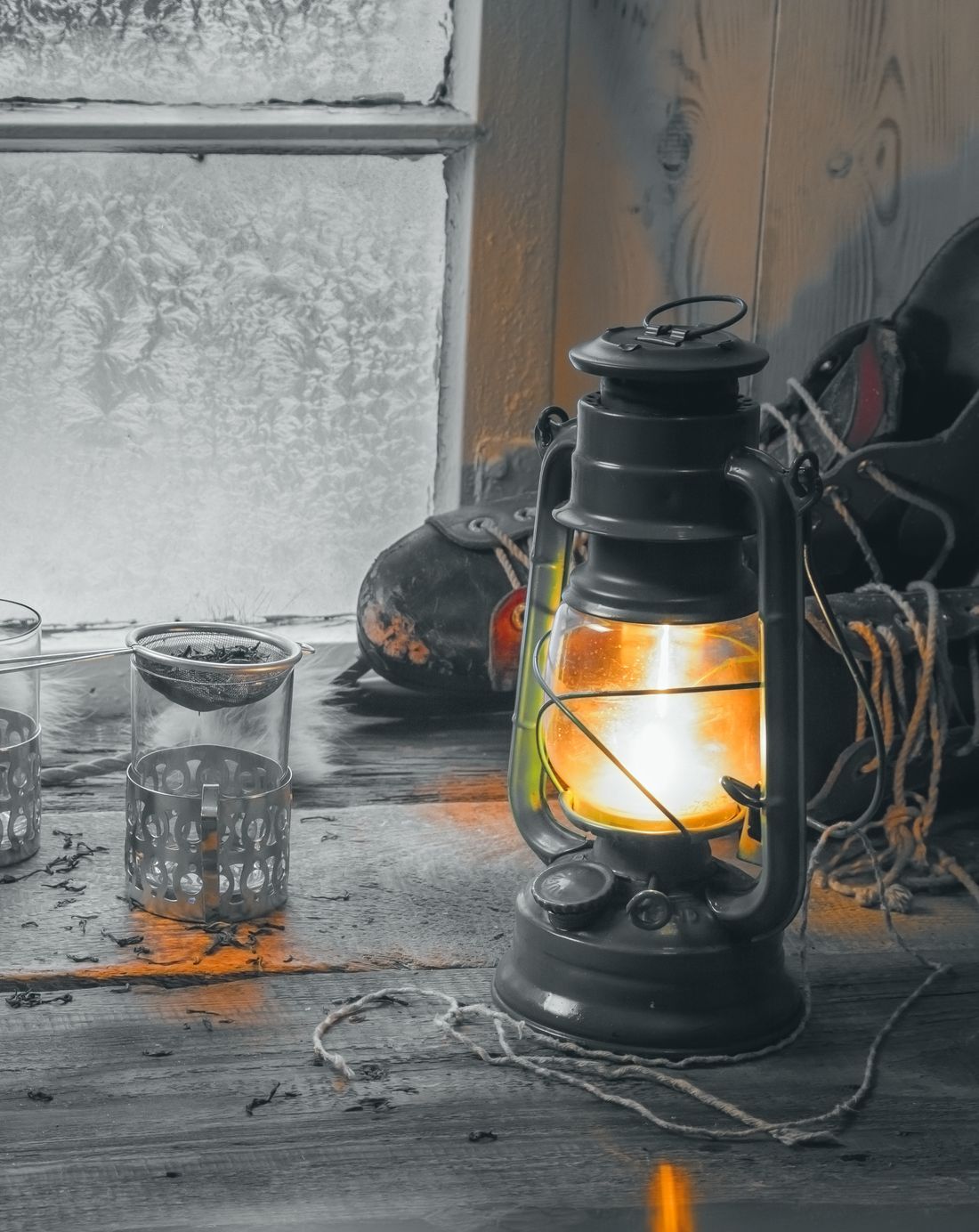 An oil lantern lit on a sill below a frosted window, printed grey: only the flame and its light on the boards keep their colour.
