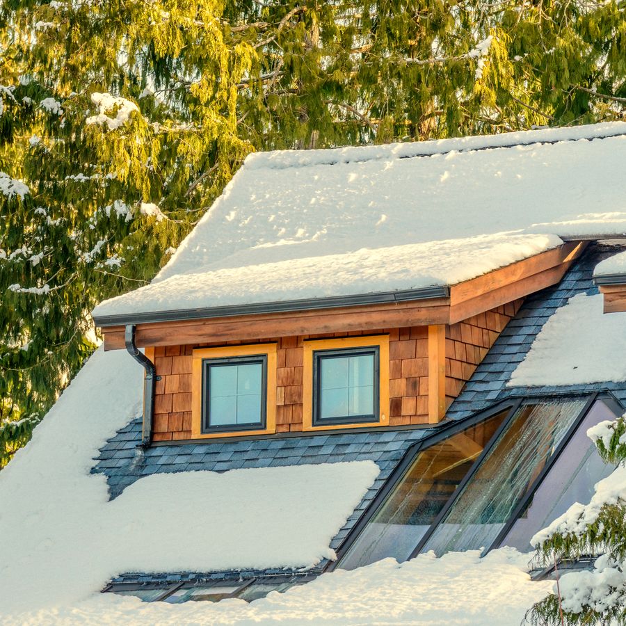 A shingled dormer with two windows on a snowy roof, in natural light.